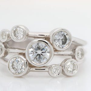 Könnte beinhalten: Ein silberner Ring mit mehreren klaren, runden Edelsteinen unterschiedlicher Größe. Der Ring hat ein einzigartiges Design mit den Edelsteinen in einer blasenartigen Anordnung, was einen funkelnden und eleganten Look erzeugt. Auf weißem Hintergrund.