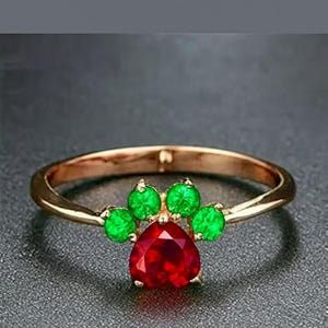 May include: A gold-colored ring featuring a paw print design. The paw print is composed of a heart-shaped, red gemstone at the center, surrounded by four green gemstones. The ring is set against a dark gray background.