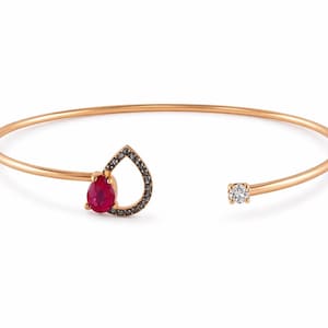 Puede incluir: Una delicada pulsera dorada con un diseño en forma de lágrima con una vibrante piedra preciosa roja y un borde de piedras más pequeñas y oscuras. Una sola piedra preciosa transparente está engastada en el extremo opuesto.