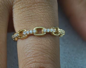 May include: A gold-colored ring with a chain link design. The ring is adorned with small, clear gemstones set along the top of the links. The ring is shown on a finger, highlighting the jewelry's details and craftsmanship.