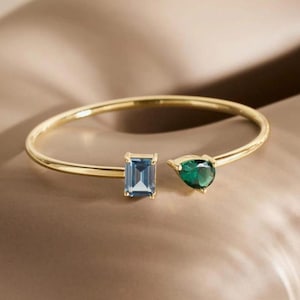 Könnte beinhalten: Ein goldfarbenes, offenes Armband mit zwei Edelsteinen. Einer ist ein rechteckiger blauer Edelstein, der andere ein birnenförmiger grüner Edelstein. Das Armband hat ein schlichtes, elegantes Design.