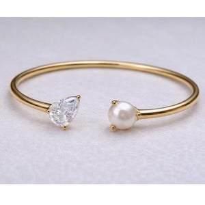 Puede incluir: Brazalete abierto dorado con un diamante en forma de pera y una perla blanca. El brazalete se presenta sobre un fondo neutro, resaltando su elegante diseño y su color dorado.