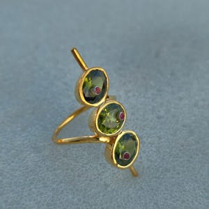 Könnte beinhalten: Ein goldener Ring mit drei olivenförmigen grünen Edelsteinen, jeder mit einem kleinen roten Akzent. Der Ring hat ein modernes Design mit einem schlanken Band und vertikalen Goldstäben, die sich von jedem Edelstein erstrecken.