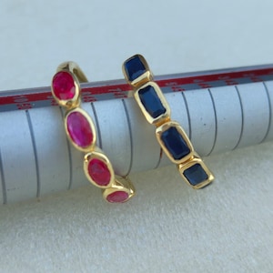 Op de afbeelding: Twee gouden ringen, één met ovale rode edelstenen en de andere met rechthoekige blauwe edelstenen. De ringen worden getoond op een ringenmeter, die hun ontwerp en kleurcontrast laat zien. De achtergrond is een neutraal off-white.