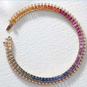 May include: A rose gold bracelet featuring a gradient of colorful gemstones, transitioning from yellow to red, purple, and blue. Small, clear stones accent the edges. The bracelet is designed with a secure clasp.