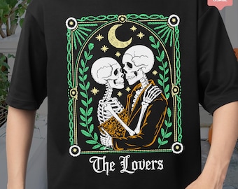 以下が含まれることがあります： 黒いTシャツ。2体の骸骨が抱き合い、三日月と星を背景に描かれています。骸骨は緑と金の装飾フレームの中にあり、下に「The Lovers」の文字があります。