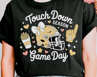 May include: Black t-shirt featuring a football-themed design. The graphic includes a gold helmet, a foam finger, hearts, stars, and the words "Touch Down Season" and "Game Day" in white script.
