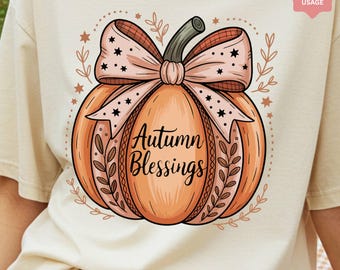 Könnte beinhalten: Beiges T-Shirt mit einer Illustration einer orangefarbenen Kürbis-Zeichnung. Der Kürbis hat einen grünen Stiel und eine Schleife mit Karomuster. Auf dem Kürbis steht "Autumn Blessings". Blätter und Sterne schmücken das Design.