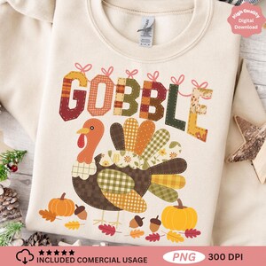 Könnte beinhalten: Cremefarbenes Sweatshirt mit einem farbenfrohen Truthahndesign und dem Wort "GOBBLE" in Patchwork-Buchstaben. Der Truthahn hat einen braunen Körper, einen orangefarbenen Kopf und gemusterte Federn. Kürbisse, Eicheln und Blätter vervollständigen das Thanksgiving-Thema.
