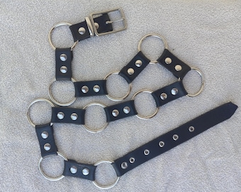 May include: Black leather harness with silver-tone metal rings and rivets. The harness features a buckle closure and an adjustable strap. The design includes multiple connecting rings and straps.