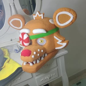 May include: A plush, orange bear head with white accents, a green band, and a red and white candy-striped eye patch. The bear has a crown, white teeth, and a yellow eye. The ears are round with white trim.