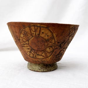 Puede incluir: Un cuenco de cerámica marrón hecho a mano con un diseño circular similar al sol tallado en el exterior. El cuenco tiene una abertura ancha y se asienta sobre una pequeña base verde claro. La superficie del cuenco tiene un aspecto texturizado.