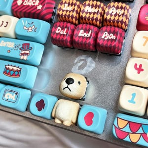 以下が含まれることがあります： さまざまな色とデザインのカスタムキーボードキーキャップのコレクション。キーキャップには、薄茶色のクマ、赤と白のダイヤモンドパターン、魔法使い、ドラムなどのイラストが描かれた水色のキーが含まれています。