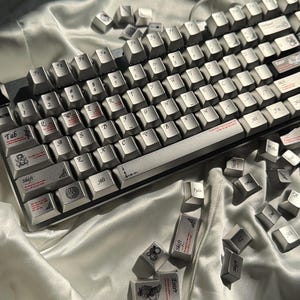 May include: A silver mechanical keyboard with keycaps scattered around. The keycaps are silver with black lettering and symbols. Some keycaps have decorative designs and text in red. The keyboard is on a white satin surface.