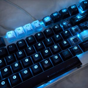 May include: A close-up shot of a backlit mechanical keyboard with black keycaps and blue LED illumination. The keys are labelled with white characters and symbols. Some keys feature unique designs. The keyboard has a silver frame and a cable.
