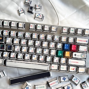 May include: A silver mechanical keyboard with custom keycaps. The keycaps feature a hand-drawn aesthetic with Japanese characters and colorful game controller icons. Some keycaps are scattered around the keyboard.