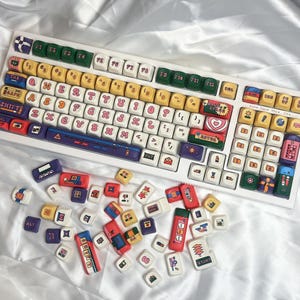 May include: A white mechanical keyboard with colorful keycaps. The keycaps feature various designs and text, including hearts, stars, and Japanese characters. Additional keycaps are scattered around the keyboard.