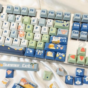 May include: A mechanical keyboard with custom keycaps in a summer theme. The keycaps are in blue, green, and white, with illustrations of a crab, watermelon, and a cat. The space bar reads "Summer Cat".