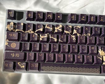 May include: A close-up of a mechanical keyboard with custom keycaps. The keycaps are a deep purple with gold accents and Japanese characters. The keyboard has a silver frame and is set against a white background.
