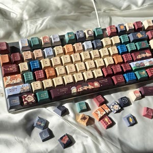 以下が含まれることがあります： 様々なデザインとイラストが施されたキーキャップを備えたカスタムメカニカルキーボード。キーキャップはベージュ、バーガンディ、ティールなど様々な色があります。一部のキーキャップにはキャラクターイラストがあり、スペースバーには「HARRY」の文字が見えます。