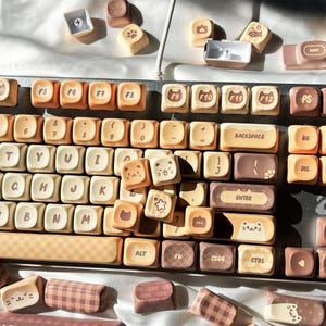 Può includere: Una tastiera personalizzata con tasti beige e marroni. I tasti presentano facce di gatto, impronte di zampe e altri simpatici disegni. La tastiera ha una varietà di tasti funzione, tra cui F5-F12, e tasti speciali come "Backspace" e "Enter".