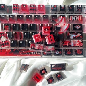Könnte beinhalten: Individuelle Tastenkappen im Thema Alice im Wunderland. Die Tastenkappen weisen rote, schwarze und weiße Designs auf, darunter Spielkarten, Rosen und Harlekinmuster. Die Tastaturtasten sind auf einer weißen Oberfläche angeordnet.