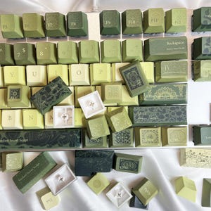 May include: A collection of green and off-white keyboard keycaps. The keycaps feature various designs and text, including "Shift", "Enter", and "Backspace". Some keycaps have floral patterns. The overall aesthetic is a mix of modern and vintage.
