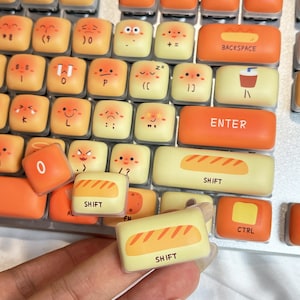 May include: Custom keyboard keycaps with a variety of emoji faces and food-themed designs. The keycaps are yellow and orange, with the word "SHIFT" on some keys. The keycaps are designed to fit a standard keyboard.