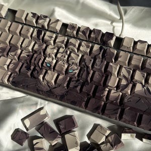 Pode incluir: Um teclado mecânico personalizado com teclas com um design de tigre. As teclas são uma mistura de cinza, castanho e branco, com uma ilustração detalhada do rosto de um tigre e olhos azuis. Algumas teclas estão espalhadas por baixo.