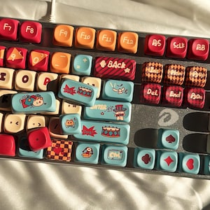 May include: A mechanical keyboard with custom keycaps in various colours. Keycaps include red, orange, and light blue. Some keycaps have cartoon illustrations and text, such as "BACK" and "Dong".