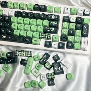 May include: A white mechanical keyboard with green and black keycaps. The keycaps feature various symbols and designs, including bats, eyes, and geometric patterns. Some keycaps are scattered around the keyboard on a white surface.