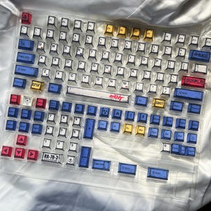 May include: A clear keyboard with custom keycaps. The keycaps feature a variety of colors, including blue, yellow, red, and white. Some keycaps have text such as "GUNPLA" and "RX-78-2". The keyboard is designed for gaming or custom builds.