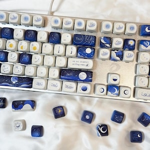 May include: A silver keyboard with custom keycaps featuring celestial designs. The keycaps are primarily white and blue, with illustrations of moons, stars, and galaxies. Some keycaps have text like "Shift" and "Menu".