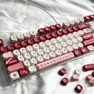 Könnte beinhalten: Eine mechanische Tastatur mit roten und weißen Tastenkappen. Die Tastenkappen haben verschiedene Designs, darunter Kirschen und Vichy-Muster. Die Tastatur hat einen silbernen Rahmen und ein weißes Kabel. Einige Tastenkappen liegen verstreut.