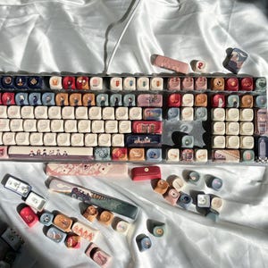 May include: A custom mechanical keyboard with a variety of colourful keycaps. The keycaps feature illustrations of people, buildings, and other designs. The keyboard is on a white surface.