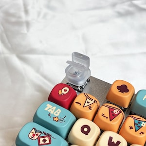 以下が含まれることがあります： カスタムキーキャップ付きキーボードのクローズアップ。キーキャップは赤、オレンジ、青など様々な色で、ユニークなデザインとテキストが特徴です。透明なプラスチック製のキーキャップも確認できます。