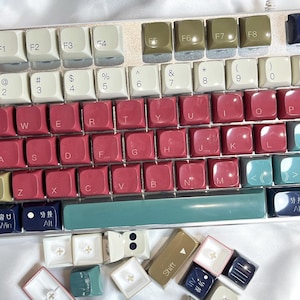 May include: A mechanical keyboard with a white, teal, and burgundy color scheme. The keycaps are a mix of white, burgundy, and teal. Several keycaps are detached and lying below the keyboard.