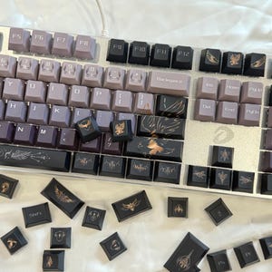 May include: A mechanical keyboard with custom keycaps. The keycaps are a mix of purple and black with gold accents. Some keycaps feature wing and emblem designs. The keyboard is on a white surface.