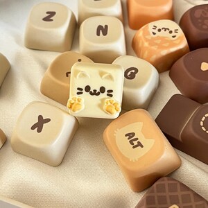 Pode incluir: Uma variedade de chocolates decorativos em forma de teclas de teclado, em vários tons de castanho e bege. Uma tecla apresenta um desenho de gato, outra tem o texto "ALT" com uma ilustração de gato. Outras teclas mostram letras.