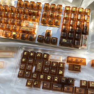 May include: A set of amber-colored mechanical keyboard keycaps with coffee-themed icons and text. The keycaps are arranged in a clear plastic tray, with additional keycaps visible on a keyboard. The text includes "COFFEE" and "CAFE".