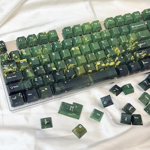 Może przedstawiać: Klawiatura mechaniczna z zielonymi i złotymi nakładkami na klawisze. Nakładki mają efekt marmuru, łącząc ciemnozielony i złoty. Klawiatura leży na białej powierzchni. Kilka nakładek jest rozrzuconych.