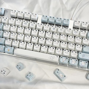 May include: A white and light blue mechanical keyboard with custom keycaps. The keycaps feature a bear face design and other cute graphics. The keyboard has a silver frame and is connected to a white cable.