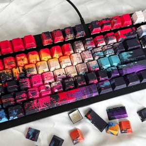 May include: A mechanical keyboard with custom keycaps. The keycaps have a vibrant, abstract design with splashes of red, orange, pink, blue, and purple. Some keycaps are removed, revealing the keyboard's switches. The keyboard is black.
