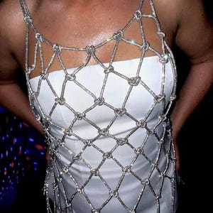 May include: A silver, sparkling, knotted chainmail top with a diamond pattern. The top has thin straps and a fringe at the bottom. The top is worn over a white dress. The background is dark.