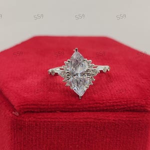 May include: A marquise-cut diamond ring with a halo of smaller stones. The ring is set on a silver band and is displayed on a red velvet box. The ring's design is elegant and eye-catching.