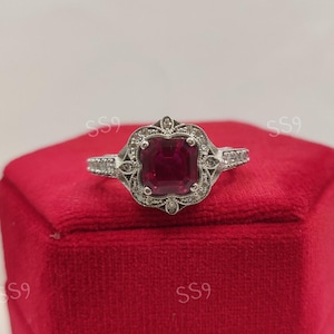 May include: A silver ring with a large, square, deep red gemstone at the center. The gemstone is surrounded by a decorative, filigree-style setting with small, clear accent stones. The ring is displayed on a red velvet surface.