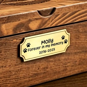 May include: A rectangular wooden box with a dark brown finish. The lid features an engraved design and a small indentation for opening. A gold-colored plaque on the front reads "Molly Forever in my memory 2016-2025" with paw prints.