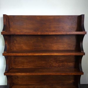 Può includere: Una mensola da parete in legno marrone scuro con tre ripiani. La mensola ha un design curvo sui lati e una parte superiore piatta. La venatura del legno è visibile e la finitura è lucida.