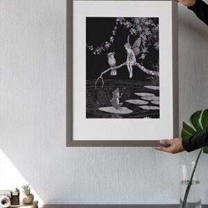 May include: A framed black and white illustration featuring a fairy, a bird, and a frog in a pond setting. The fairy is perched on a branch with the bird. The frog sits on a lily pad. The artwork is held by a person.
