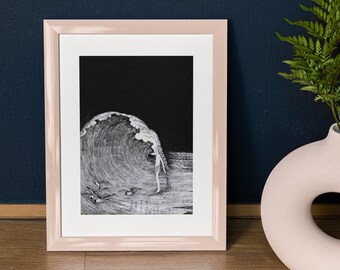 May include: A framed black and white art print depicts a surfer riding a large wave. The artwork is in a light pink frame, set against a dark blue wall. A white vase with a circular opening and green fern adds a decorative touch.